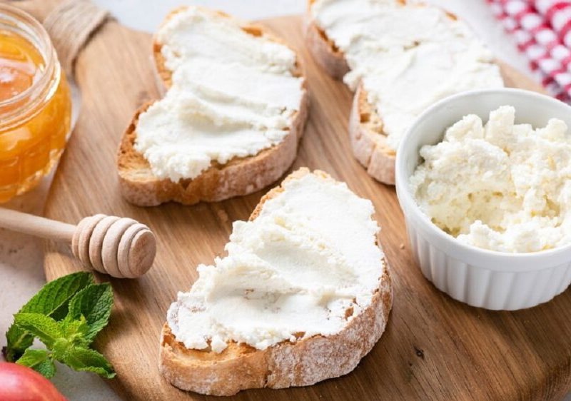 auf einem Tisch liegen drei Weißbrotscheiben mit Frischkäse. Daneben ein Glas Honig, eine Nektatrine und ein Schälchen mit weißem Frischkäse. Es handelt sich um Ricotta, einen italienischen Käse.