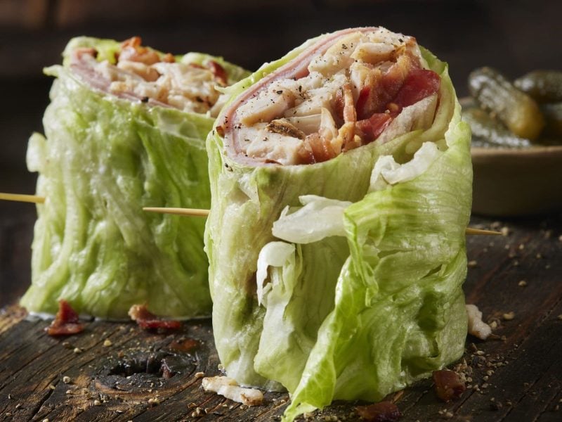 Salat-Wrap mit Pute und Bacon auf einem Holzbrett. Im Hintergrund eine Schale mit Cornichons.