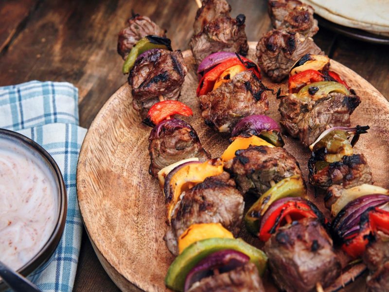 Türkische Shish Kebab mit Tomaten, Zwiebel und Paprika türkische Lammspieße auf rundem Holzbrett auf Holztisch. Daneben Schale mit hellem Dip auf blau-weißem Tuch. Im Hintergrund Fladenbrot. Aufnahme aus der Vogelperspektive.