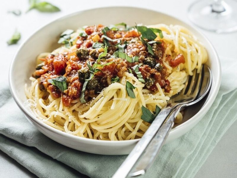 auf einem weißen Untergrund liegt ein graues, festes Tuch. Auf diesem steht ein weißer Teller. In dem Telle sind Spaghetti mit Tomaten-Sardellen-Sauce. In dem Teller liegen ein Löffel und eine Gabel.