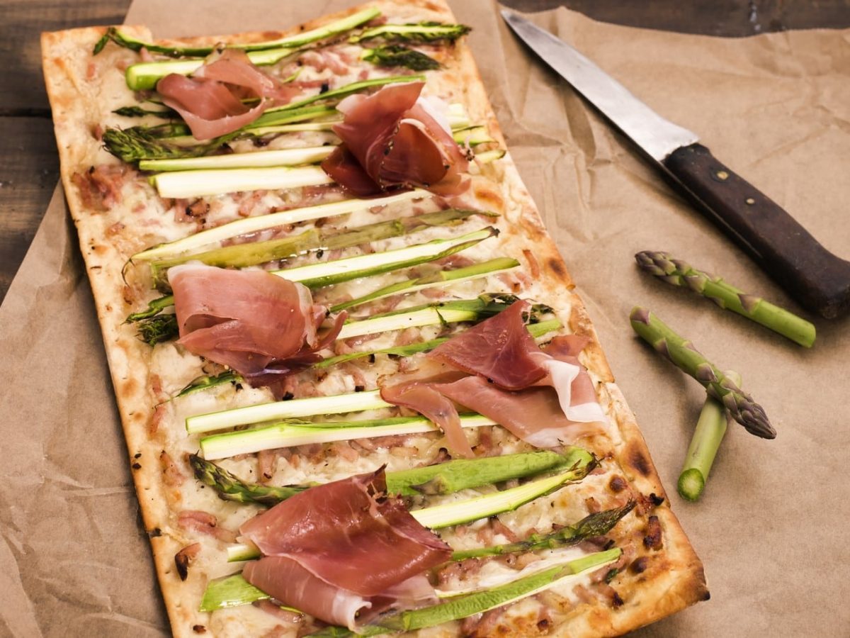 Spargel-Flammkuchen mit Serranoschinken auf einem Backpapier. Daneben etwas Spargel und ein Messer.