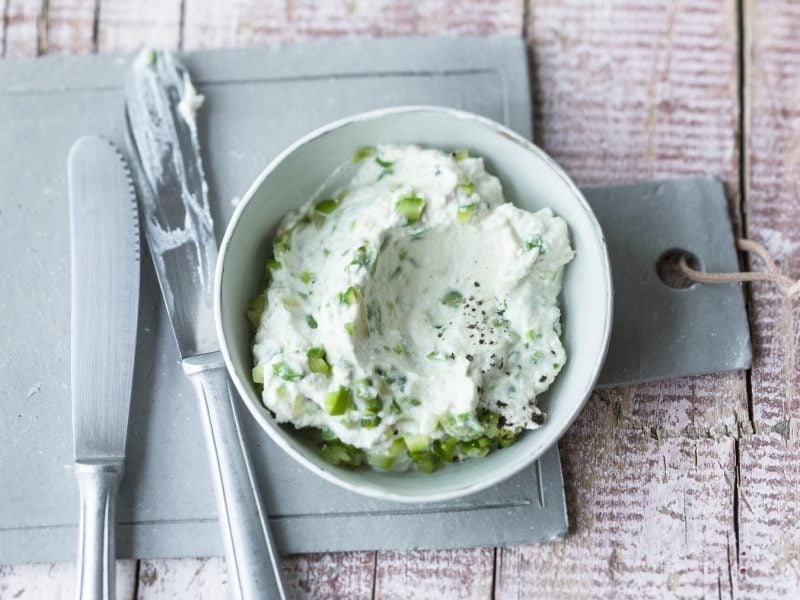 Spargel-Ricotta-Creme in einer weißen Schale auf einem Schneidebrett. Daneben zwei Messer. Das Ganze auf einem hölzernen Untergrund.