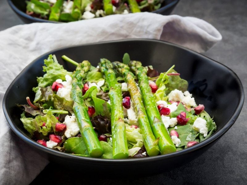 Gemischter grüner Salat mit Feta, Granatapfelkernen und gebratenem grünen Spargel in einer schwarzen Schale auf dunklem Untergrund.