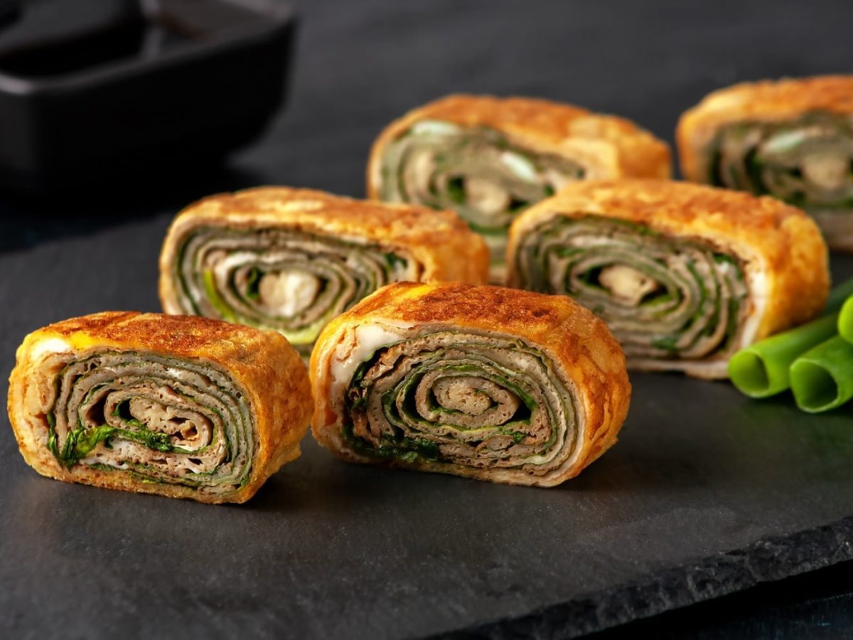 Tamagoyaki mit Pilzen und Lauch in 6 Stücke geschnitten auf einer schwarzen Schieferplatte. Im Hintergrund ein schwarzes Schälchen mit Sojasauce und Essstäbchen.