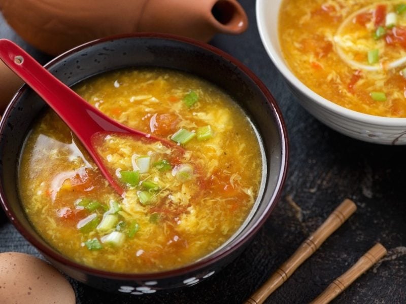 Egg-Drop-Suppe mit Tomate in einer schwarzen Schüssel auf einer schwarzen Steinoberfläche neben ein paar Eier und Stäbchen