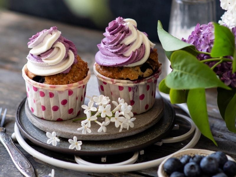 Zwei vegane Heidelbeer-Cupcakes auf einem Teller, daneben stehen frische Blumen und eine Schale mit Blaubeeren.