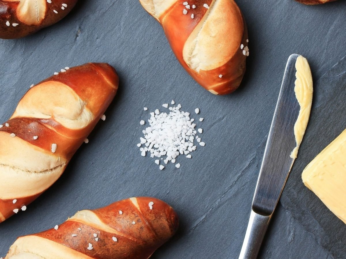 Vegane Laugenstangen, ein Buttermesser, Butter und Salz auf grauem Untergrund in der Draufsicht.