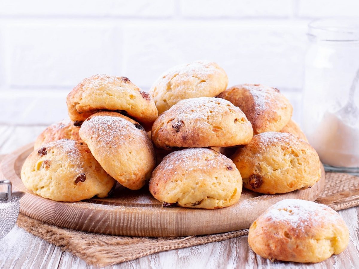 Ein Haufen veganer Rosinenbrötchen auf einem runden Holzbrett in hellem Setting, daneben ein Sieb und ein Glas Puderzucker.