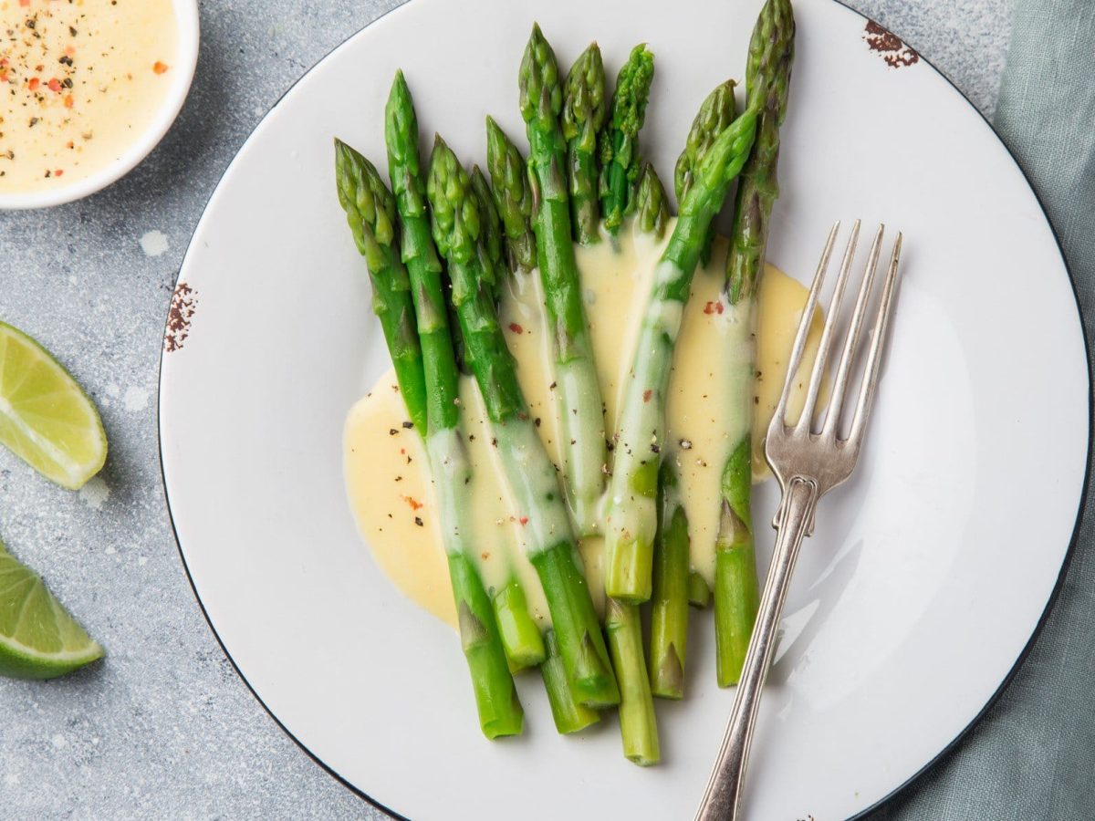 Ein Teller grüner Spargel mit veganer Sauce hollandaise und einer Gabel in der Draufsicht, daneben Limettenschnitze und eine Schale der Sauce.