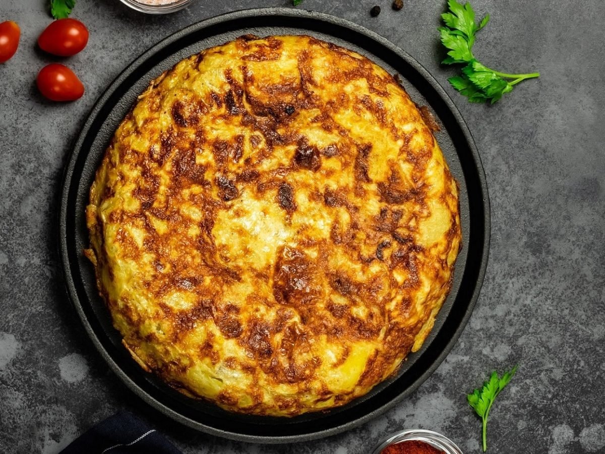 Eine vegane spanische Tortilla auf dunklem Untergrund in der Draufsicht, daneben liegen Kirschtomaten und frische Kräuter.