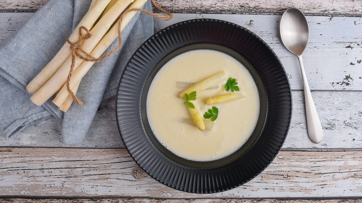 Ein schwarzer Teller vegane Spargelcremesuppe mit Spargelspitzen und etwas Petersilie auf Holz in der Draufsicht, daneben ein Löffel und frische Spargelstangen auf einem Geschirrtuch.