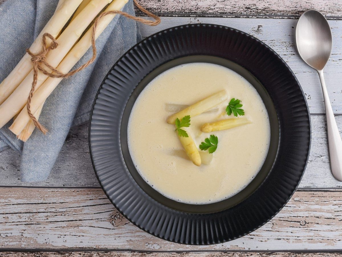 Ein schwarzer Teller vegane Spargelcremesuppe mit Spargelspitzen und etwas Petersilie auf Holz in der Draufsicht, daneben ein Löffel und frische Spargelstangen auf einem Geschirrtuch.