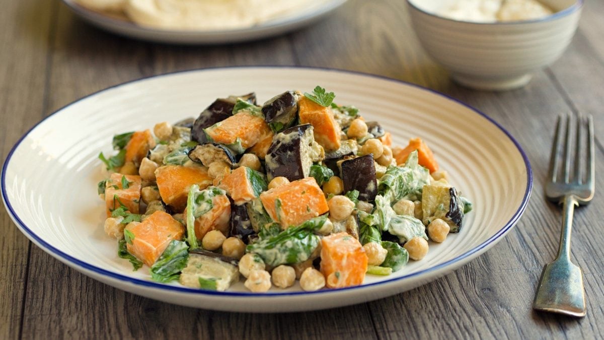 Ein Teller Veganer Kichererbsensalat mit Süßkartoffel, Aubergine und Spinat, daneben liegt eine Gabel.