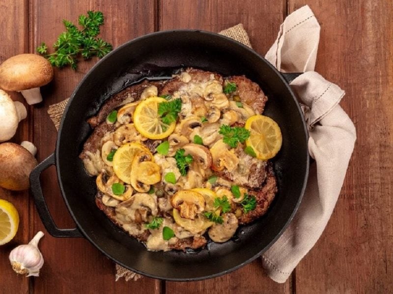 In einer Pfanne, die auf einem Tisch steht, sind Zitronen-Schnitzel. Neben der Pfanne liegen Pilze, Petersilie, Knoblauch und ein Tuch.