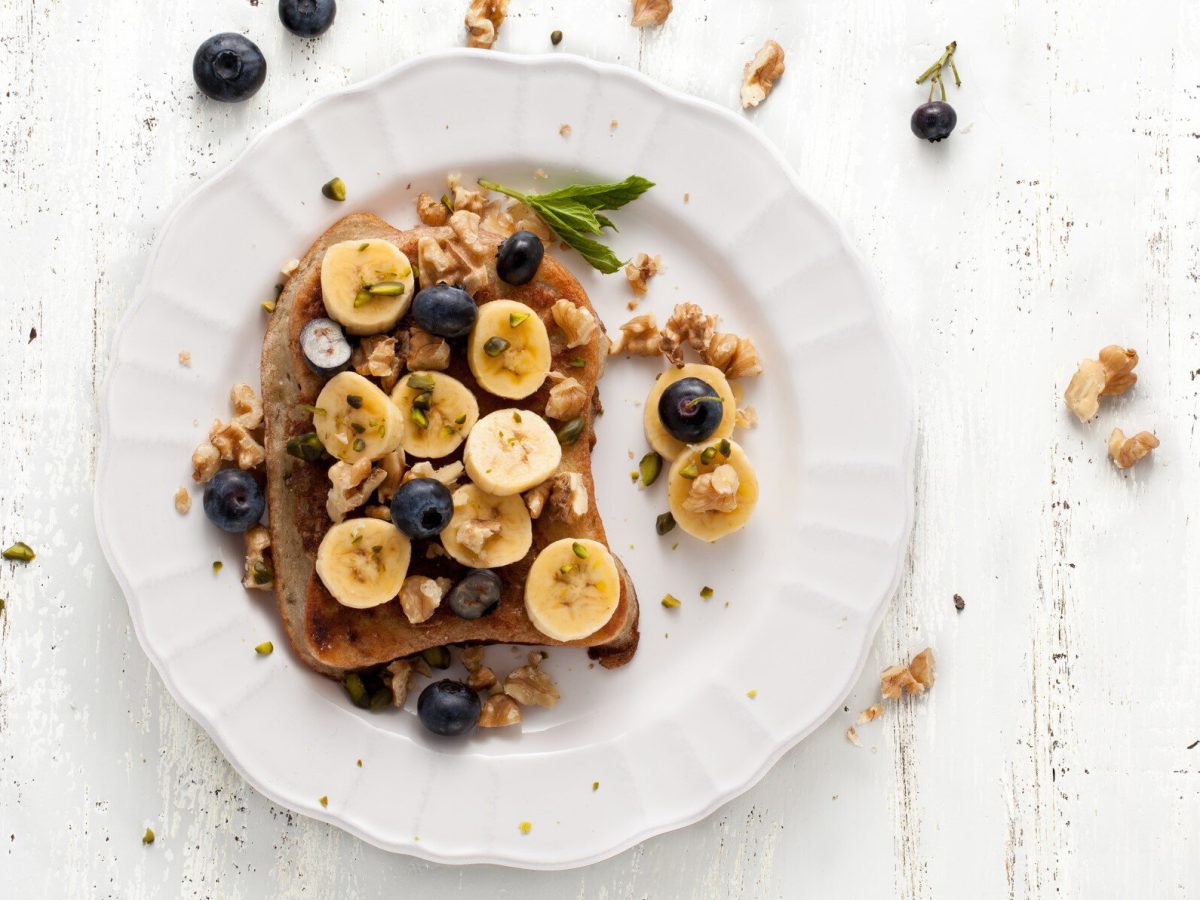 Arme Ritter mit Banane, Blaubeeren und Walnuss auf weißem Teller.