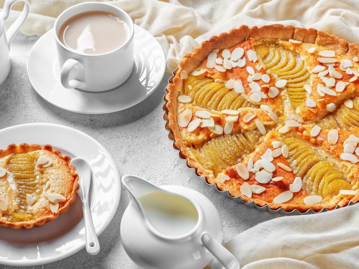 Birnen-Flan-Tarte, bestreut mit Mandeln, angeschnitten serviert auf einem mit weißen Tüchern ausgedeckten Kaffetisch.