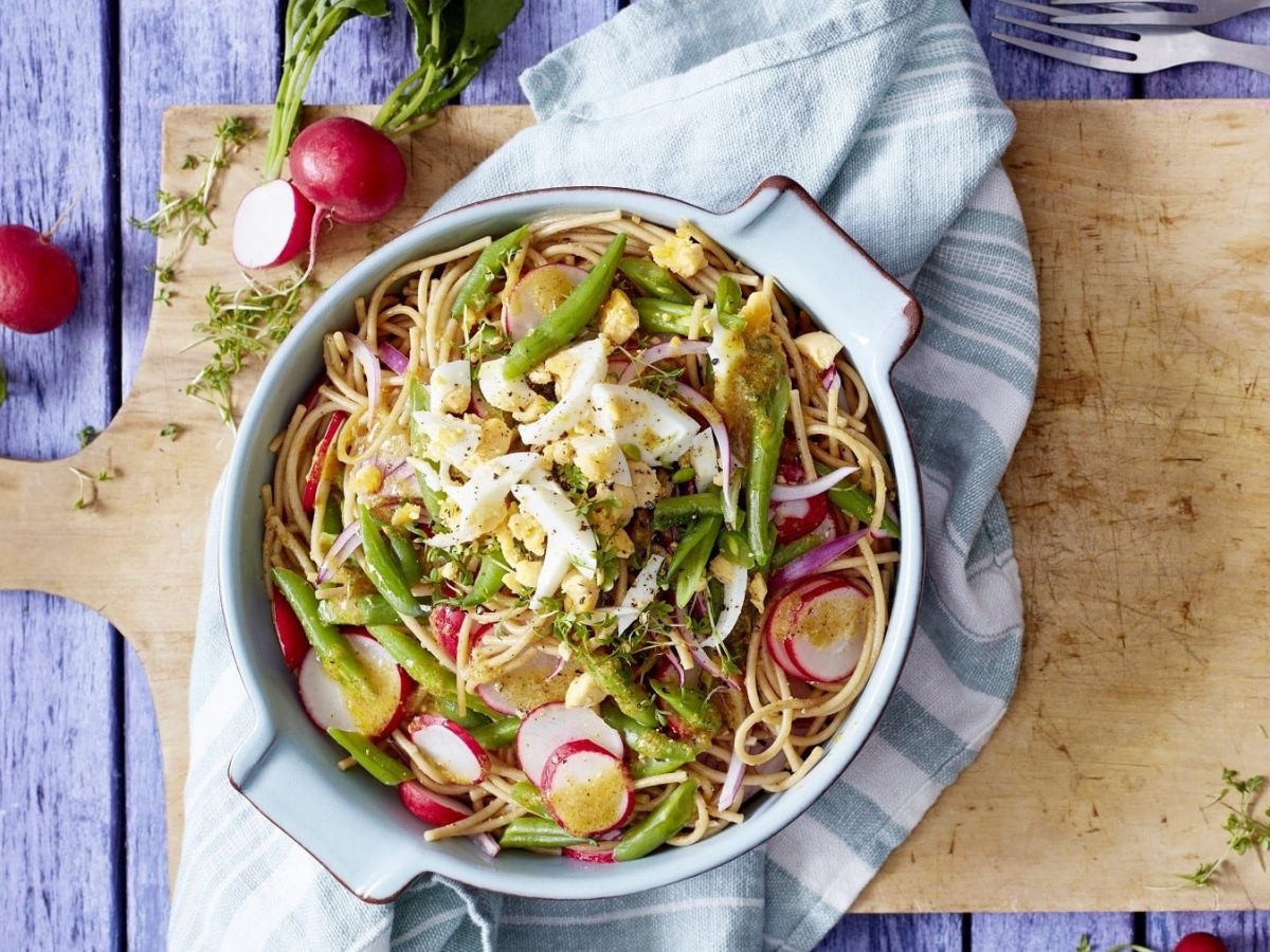 Bunter Bohnen-Nudel-Salat mit Ei, grünen Bohnen, Radieschen, roten Zwiebeln und Weißweindressing, serviert in einer großen Auflaufform auf einem rustikalen Holzbrett.