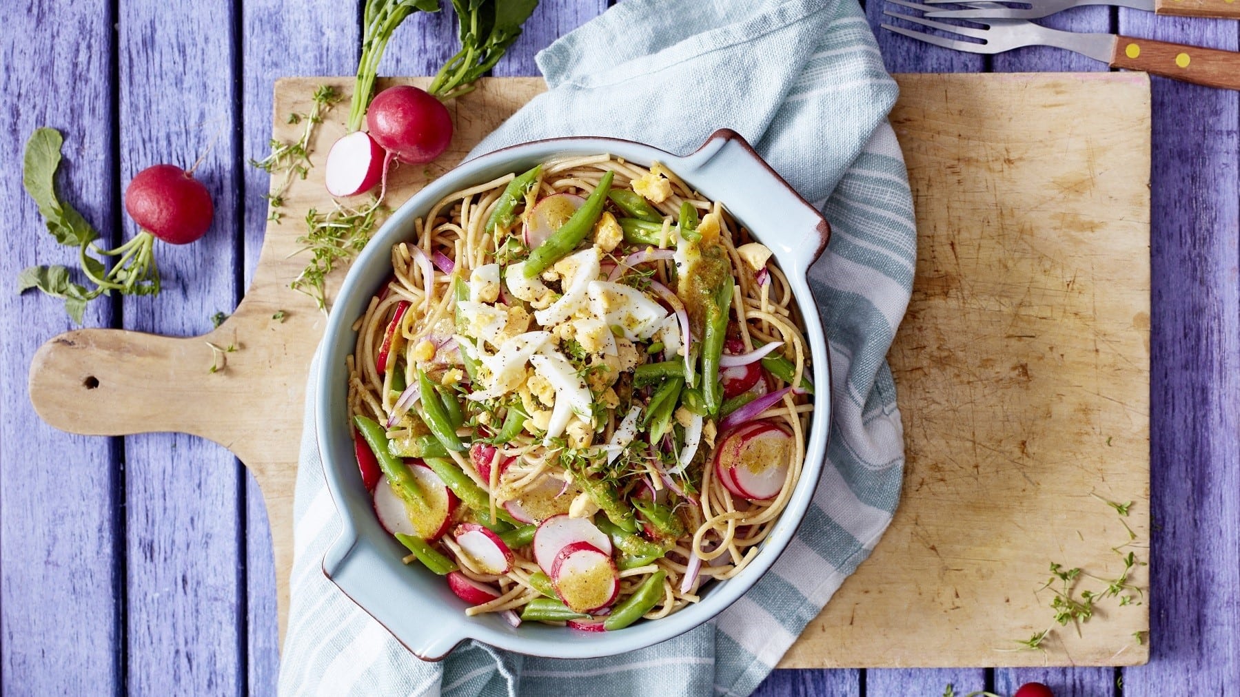 Bunter Bohnen-Nudel-Salat mit Ei, grünen Bohnen, Radieschen, roten Zwiebeln und Weißweindressing, serviert in einer großen Auflaufform auf einem rustikalen Holzbrett.