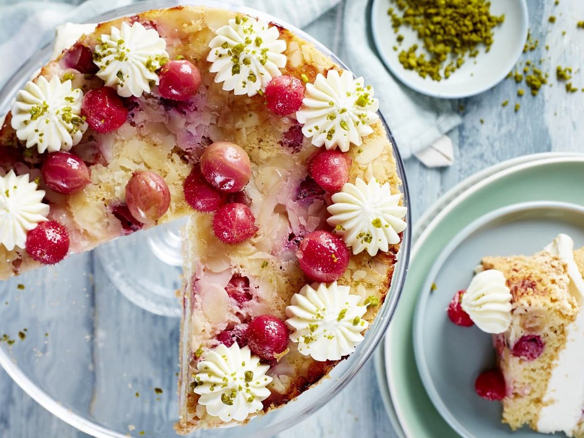 Auf einem Tisch steht eine Stachelbeer-Torte. Sie ist mit frischen Stachelbeeren und Mandelblättchen dekoriert. Am Rand wurden Tuffs aus Creme aufgetragen.