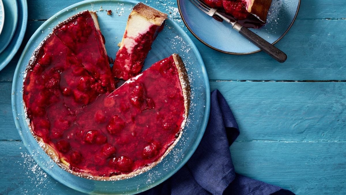 Auf einem Teller liegt einNew York Cheesecake mit Himbeeren. Er ist angeschnitten und man erkennt die fluffige Cremefüllung.