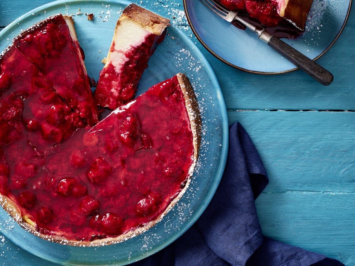 Auf einem Teller liegt einNew York Cheesecake mit Himbeeren. Er ist angeschnitten und man erkennt die fluffige Cremefüllung.