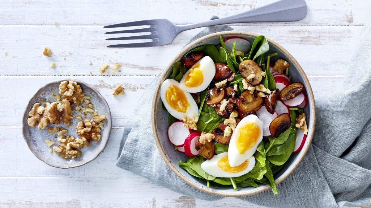 Frühlings-Bowl mit Ei, Babyspinat, Radieschen, Pilzen und angerösteten Walnüssen und gekochtem Ei, serviert auf einem hellen Tisch in einer Schale.