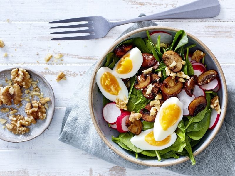 Frühlings-Bowl mit Ei, Babyspinat, Radieschen, Pilzen und angerösteten Walnüssen und gekochtem Ei, serviert auf einem hellen Tisch in einer Schale.