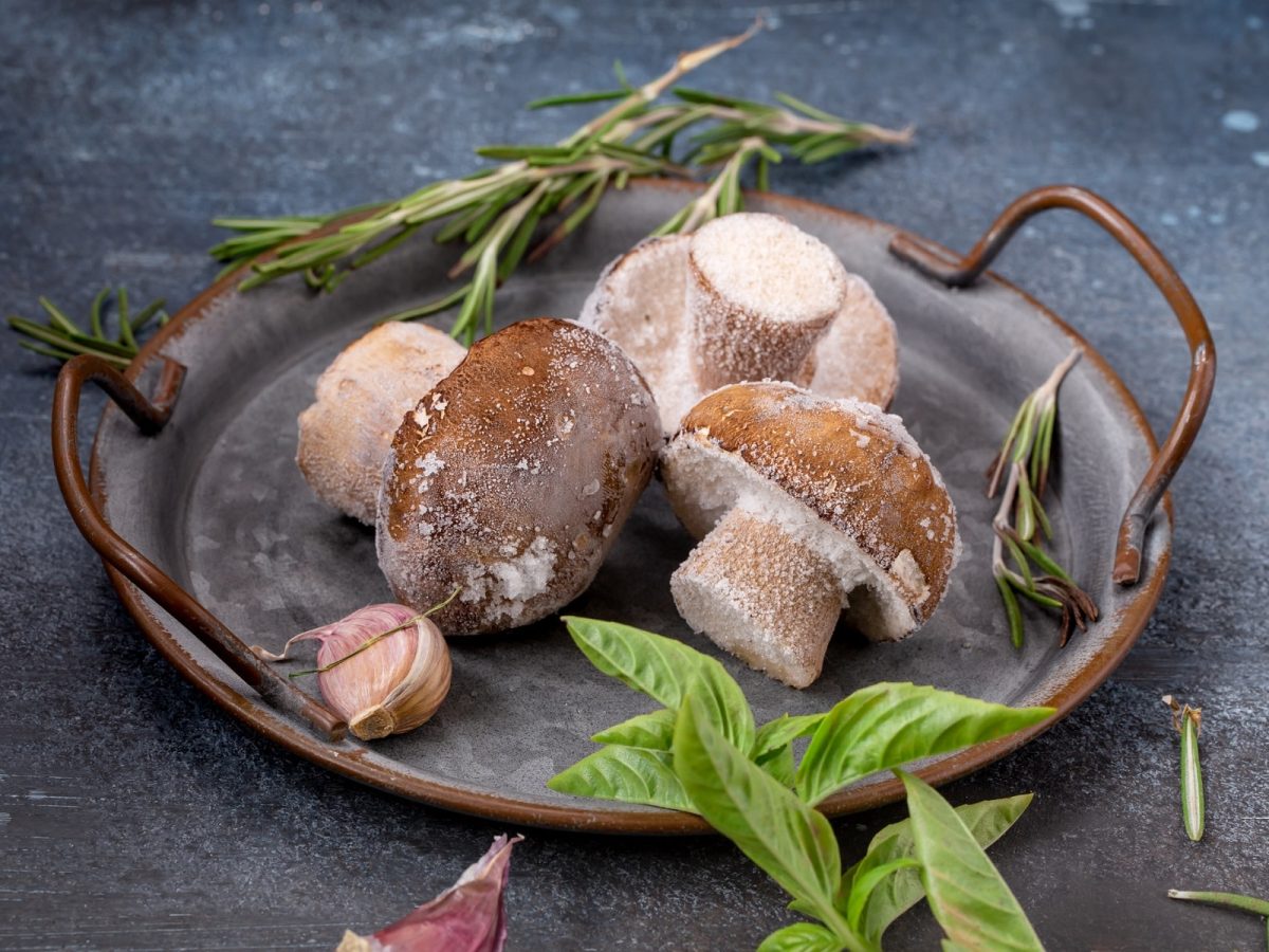 Drei gefrorene Pilze nebst Knoblauch und Kräutern in einer Metallschale.