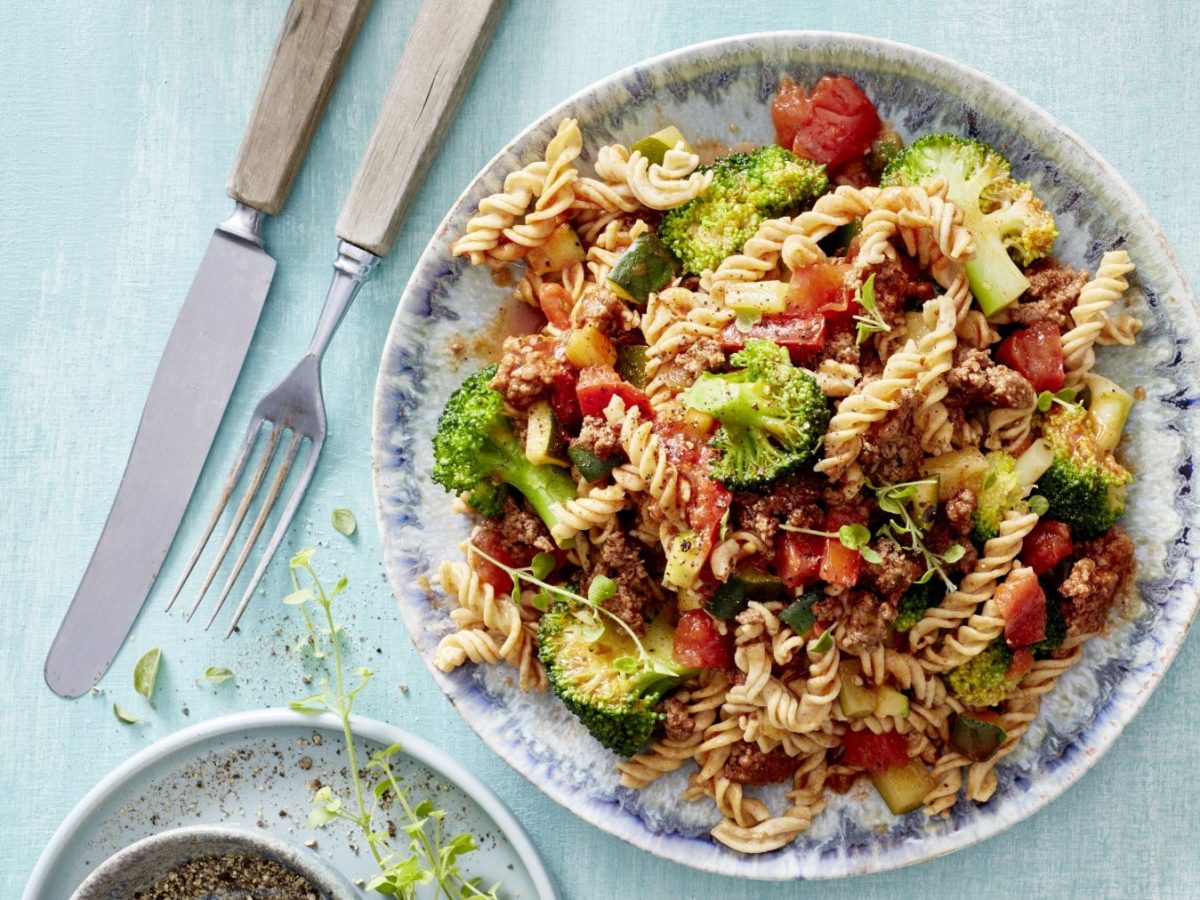 Nudelsalat mit Hack und Gemüse auf einem gemusterten Teller. Daneben Gewürze und Besteck.