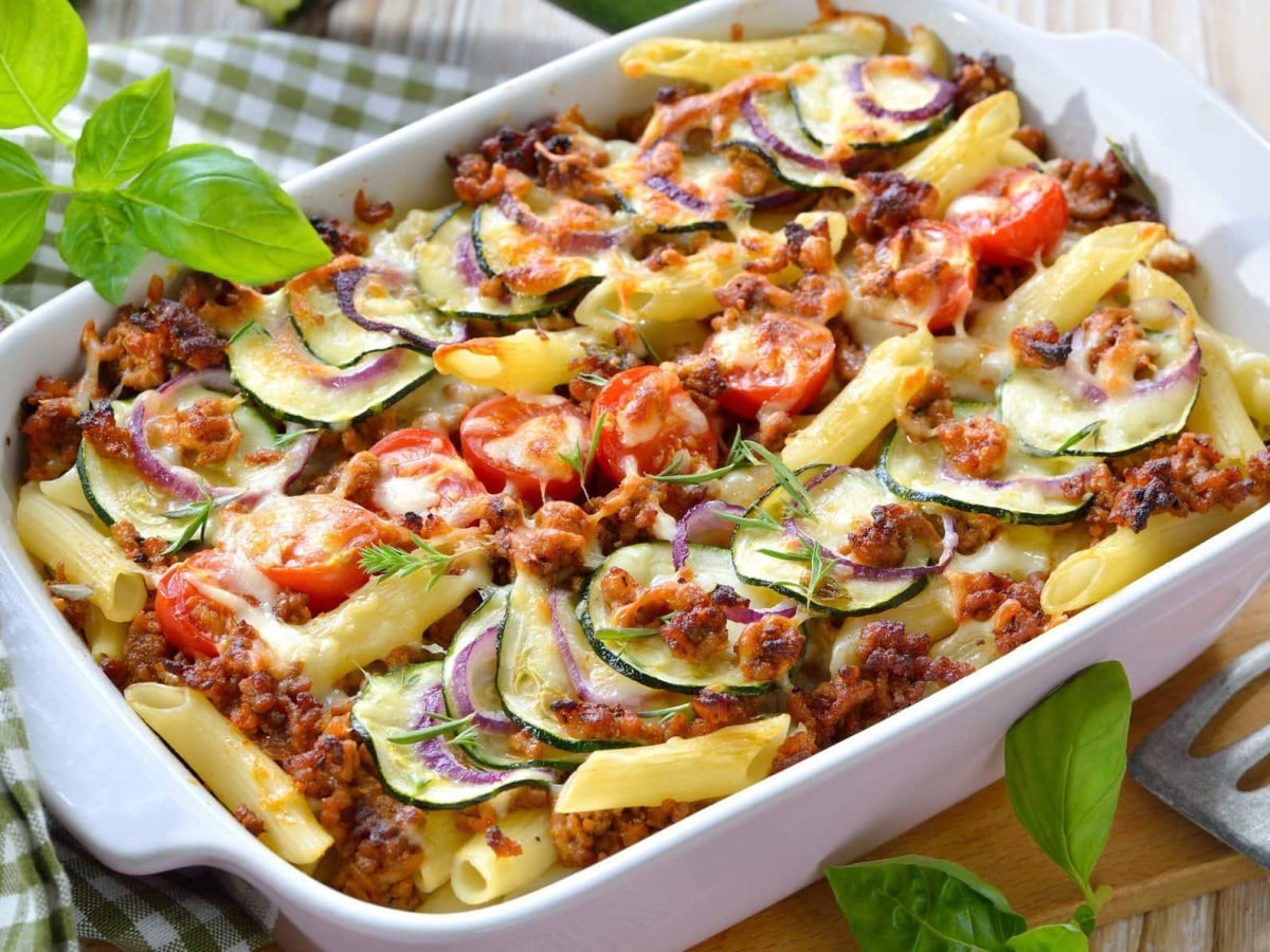 Pasta al forno mit Hackfleisch und Zucchini in weißer Auflaufform. Daneben liegt ein weiß-grün-kariertes Tischtuch