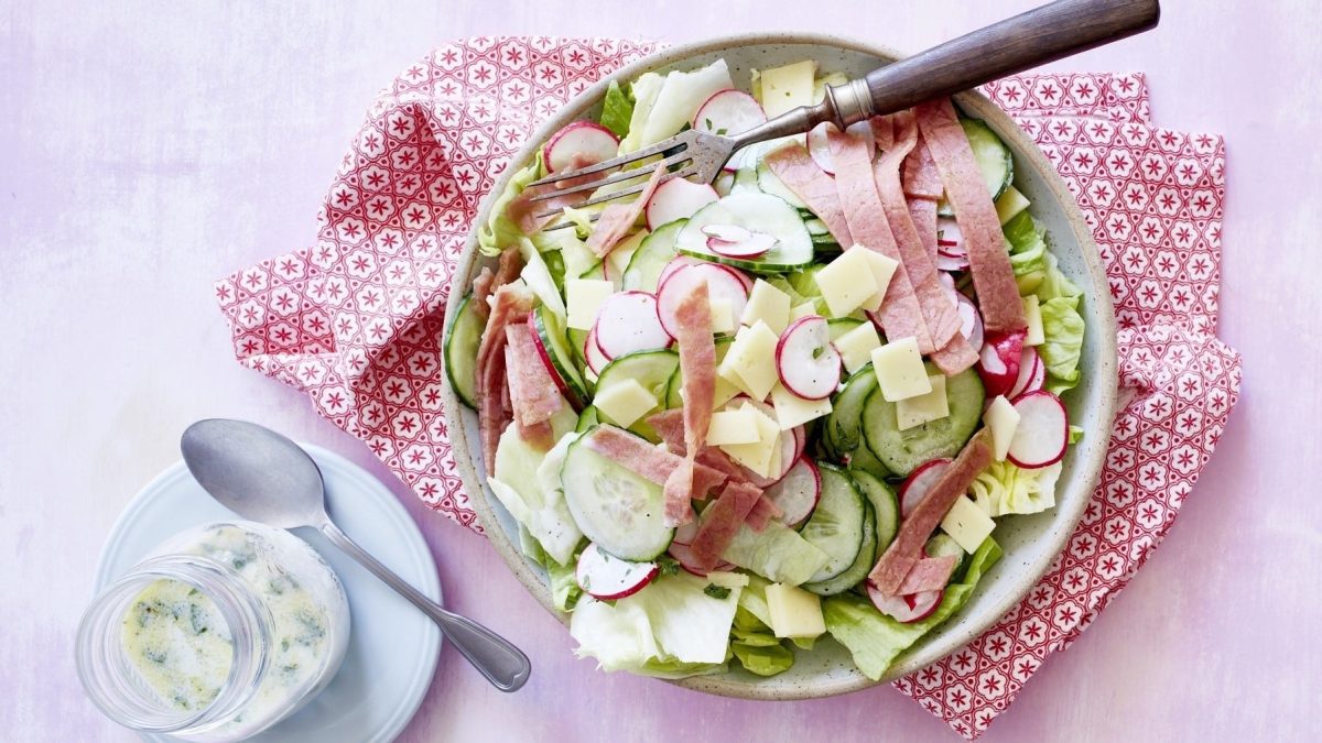 Salat mit Käse und Radieschen, garniert mit frischer Petersilie, einem Dressing aus Molke und Zitrone und in Scheiben geschnittenem Corned Beef. Das ganze ist auf einer Basis von Gurke und Salat drappiert und in einer hellen Schale auf rosanem Untergrund serviert.