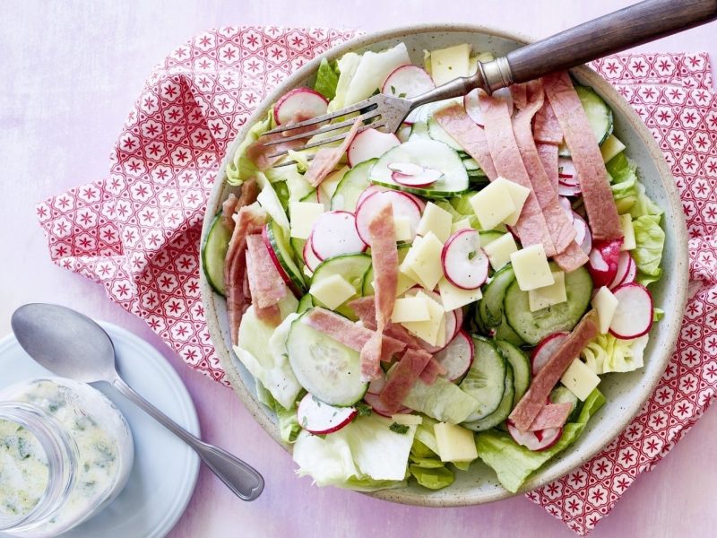 Salat mit Käse und Radieschen, garniert mit frischer Petersilie, einem Dressing aus Molke und Zitrone und in Scheiben geschnittenem Corned Beef. Das ganze ist auf einer Basis von Gurke und Salat drappiert und in einer hellen Schale auf rosanem Untergrund serviert.