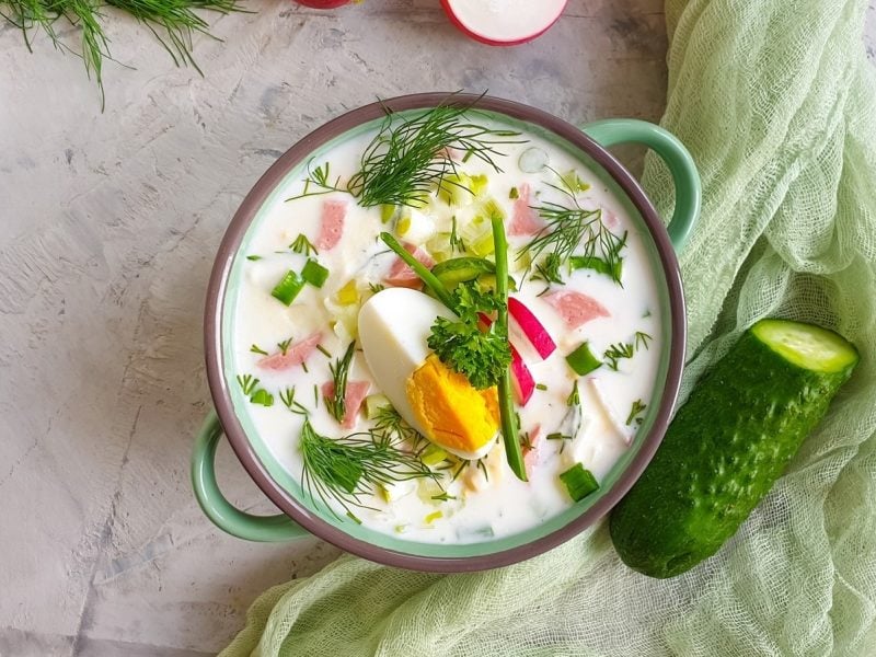 In einer Schüssel ist eine kalte Gurkensuppe angerichtet. Daneben liegt eine Gurke.