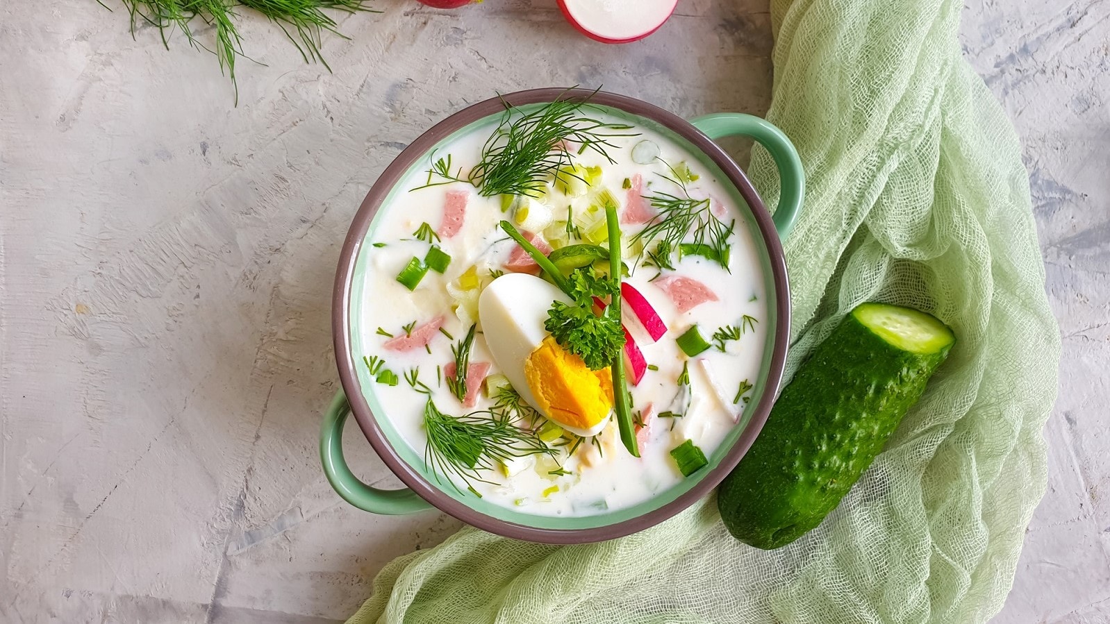 In einer Schüssel ist eine kalte Gurkensuppe angerichtet. Daneben liegt eine Gurke.