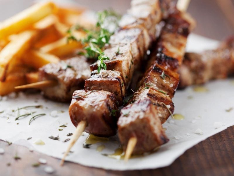 Souvlaki-Spieße mit Pommes frites auf Papier, angerichtet auf Holzuntergrund.