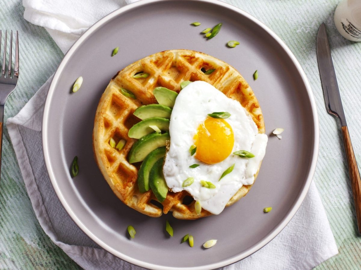 Waffel mit Avocado und Spiegelei auf einem grauen Teller. Daneben Besteck und ein Salzstreuer.