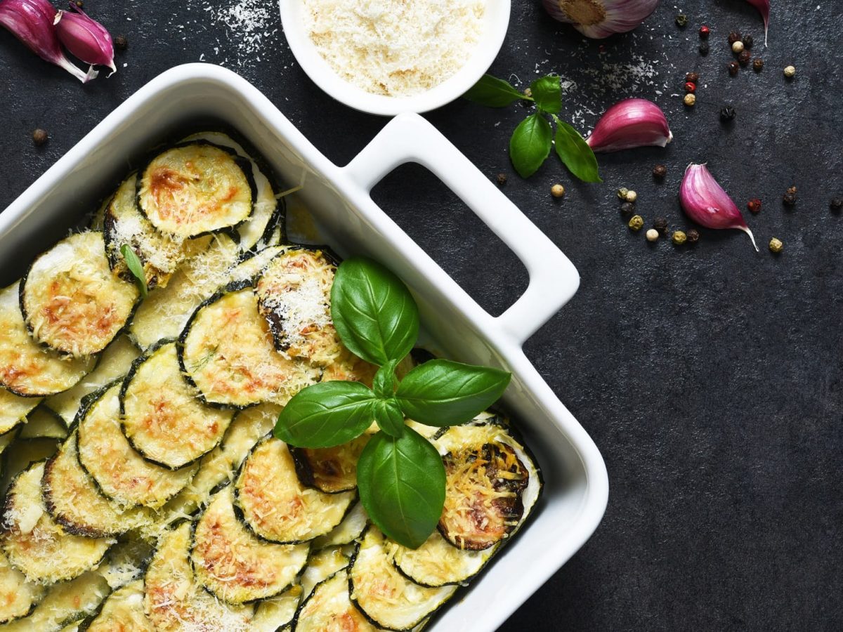 Zucchini-Gratin in weißer Form auf Schieferuntergrund. Daneben liegen Gewürze und Knoblauch