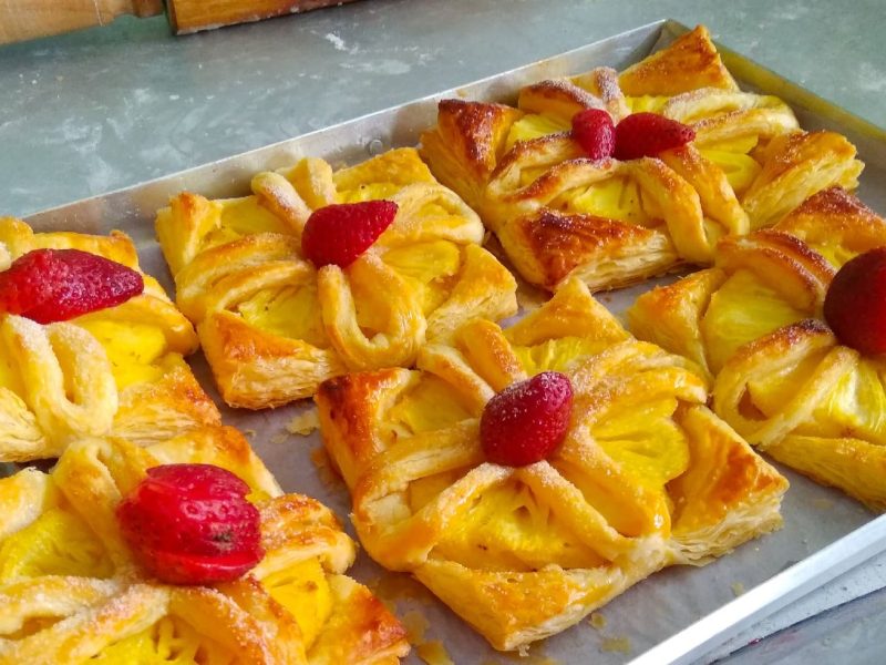 Sechs Ananas-Erdbeer Puffs auf einem Backblech mit Backpapier auf einem steinuntergrund und einem Küchenmesser.