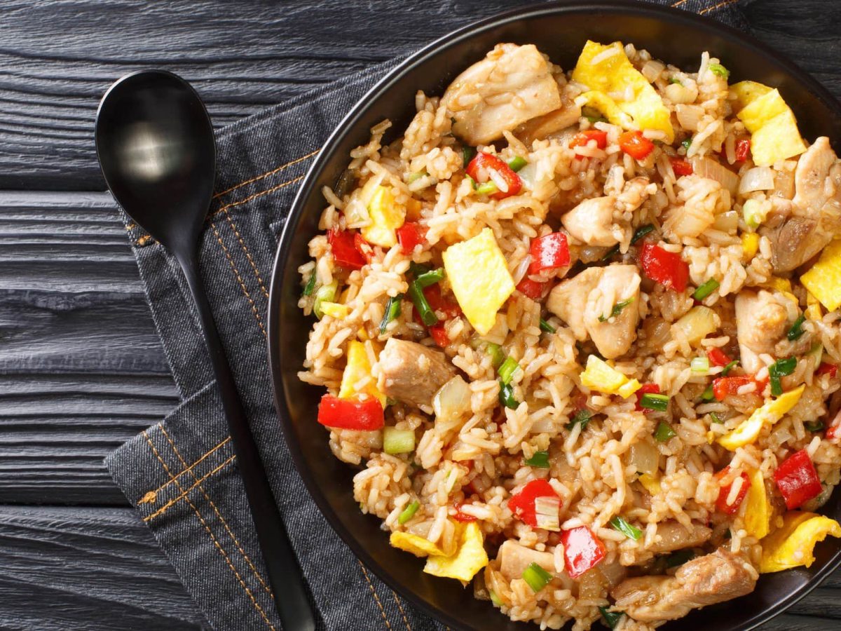 Arroz Chaufa peruanischer Reis mit Huhn in schwarzer Gusspfanne auf Jeanstuch und dunklem Holz. Daneben schwarzer Löffel. Aufnahme aus der Vogelperspektive.