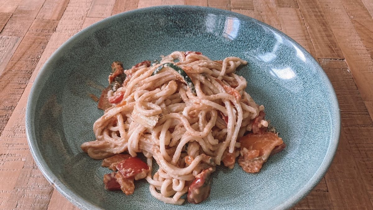 Ein Teller asiatische Pasta mit Miso-Gemüse und Erdnusssauce auf einem blau-grünen Teller, der af einem Holztisch steht.