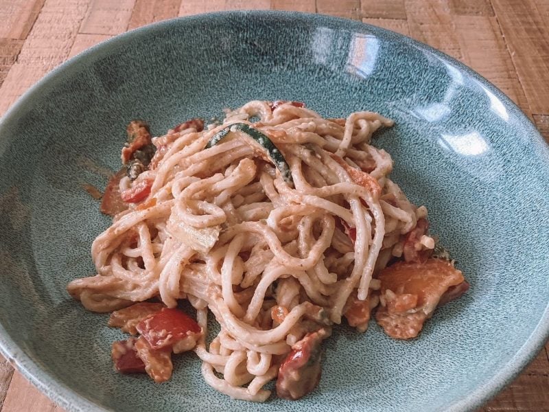 Ein Teller asiatische Pasta mit Miso-Gemüse und Erdnusssauce auf einem blau-grünen Teller, der af einem Holztisch steht.