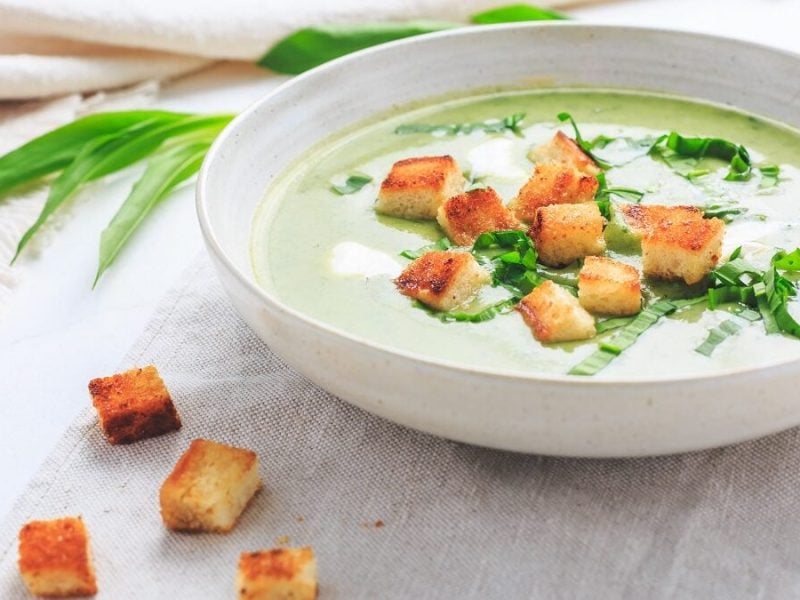 Ein Teller mit einer Bärlauch-Blumenkohl-Suppe mit Croûtons steht auf einem Untergrund aus Holz. Daneben lieben Bärlauch-Blätter und Brotwürfel.