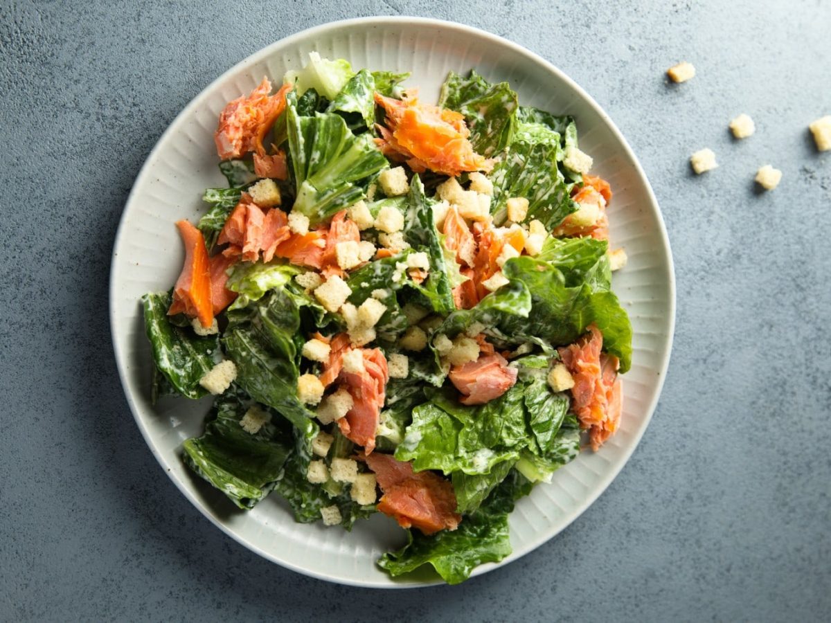 Caesar Salat mit Lachs aus einem weißen Teller. Daneben liegen Croutons.