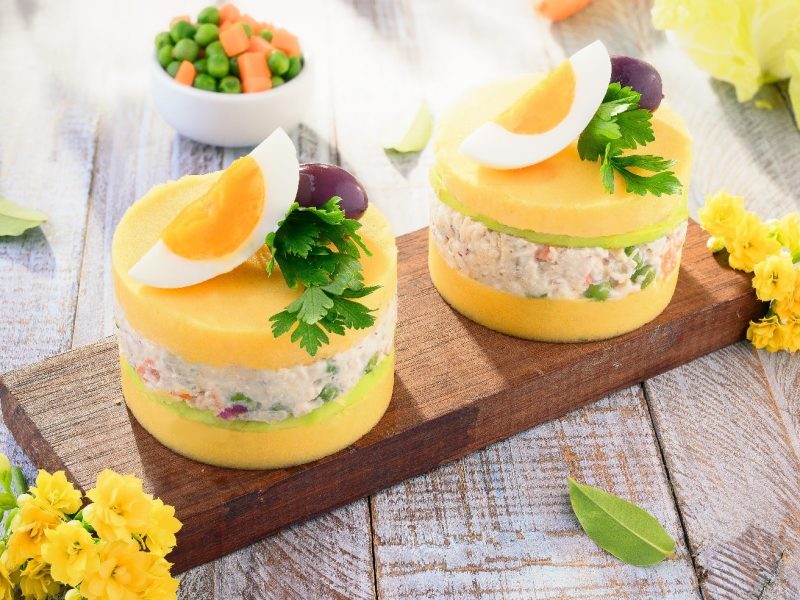 Zwei Portionen Causa limena auf einem Holzbrett mit Blüten und Gemüse drum herum auf einem Holztisch.