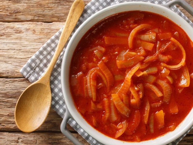 Cebolada portugiesische Zwiebelsauce in emailliertem Topf mit weißem Rand auf karierter Stoffserviette auf Holztisch. Daneben Holzlöffel. Aufnahme aus der Vogelperspektive.