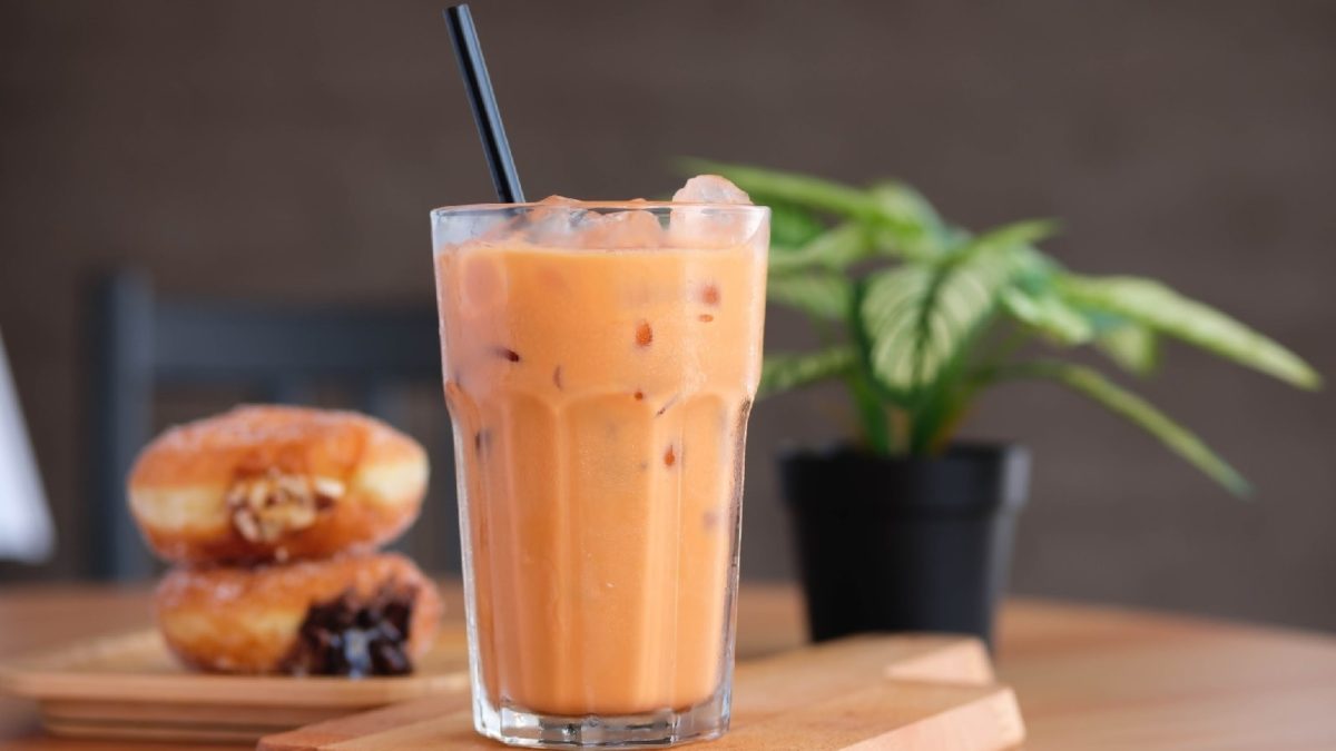 Ein Glas mit orangenem Thai-Eistee auf einem Holztisch vor einer grauen Wand