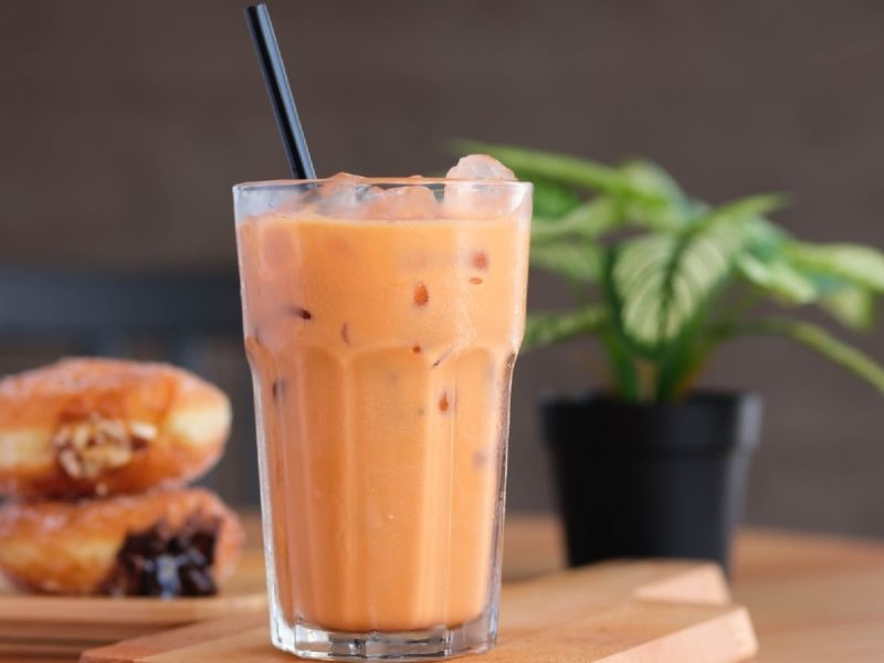 Ein Glas mit orangenem Thai-Eistee auf einem Holztisch vor einer grauen Wand