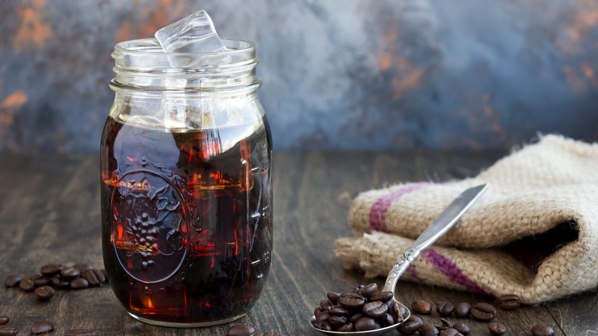 Ein Glas Cold Brew Coffee mit Eiswürfeln, daneben ein Löffel mit Kaffeebohnen und ein Geschirrtuch.