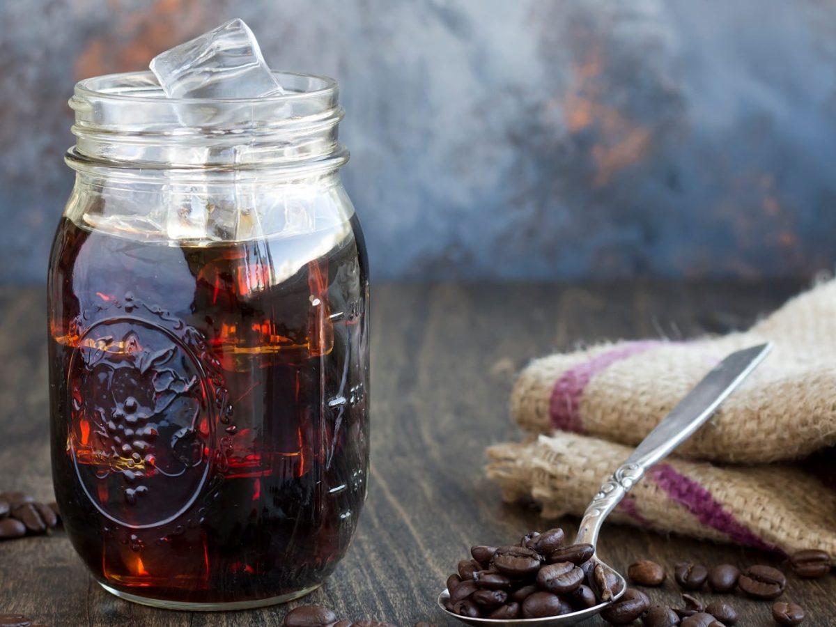 Ein Glas Cold Brew Coffee mit Eiswürfeln, daneben ein Löffel mit Kaffeebohnen und ein Geschirrtuch.