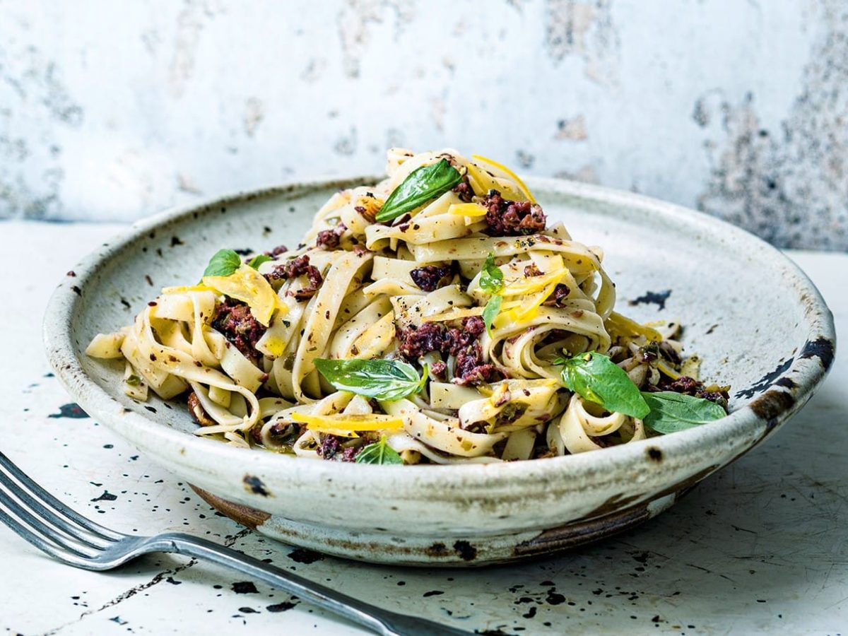 Ein Teller Tagliatelle mit Oliventapenade und Basilikum, daneben liegt eine Gabel, in rustikalem Setting. Das Rezept stammt aus dem Kochbuch "Deftig vegan mediterran".